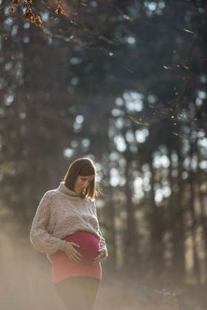Young woman in late pregnancy touching her swollen belly looking at it lovingly while standing outside in a forest with sunlight around her.の写真素材