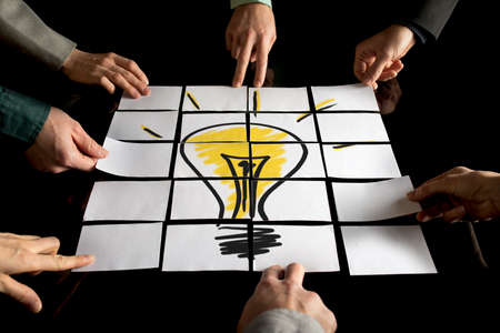 Brainstorming and teamwork concept with a team of seven businesspeople arranging white papers forming a yellow light bulb on a black table.の写真素材