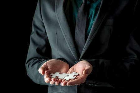 Businessman in black suit and tie holding two matching puzzle pieces, isolated on black background with copy space.の写真素材