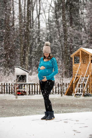 Pregnant woman holding her belly standing outside in winter hat and blue sweater under snowfall in rural area.の写真素材