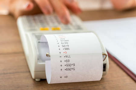 Woman using a manual adding machine to do the accounts and balance the books in the office or home, closeup low angle view with focus to the figures on the paper strip in a financial concept.の写真素材