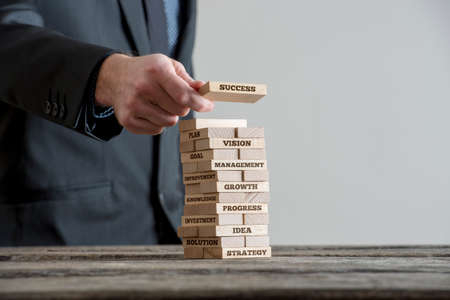 Businessman in black suite building tower of wooden domino bricks with motivational business concept signs about company building strategy. Close-up on wooden table with grey wall copy space.の写真素材