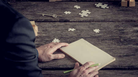Retro image of blank notebook with copy space in front of incognito businessman cropped image, hands with pencil over old rough wooden table surface with puzzle pieces and key.の写真素材