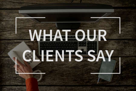What our clients say text over top view image of hand reaching for phone seated in front of computer keyboard, monitor and notepad.の写真素材