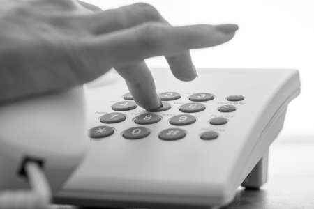 Monochrome image of female hand dialing a telephone number on a white landline phone.の写真素材
