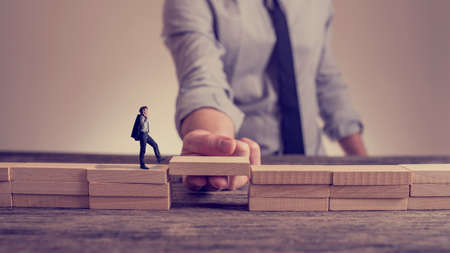 Man solving problems by building bridge with wooden block to span a gap for little businessman walking across in a conceptual retro toned image.の写真素材