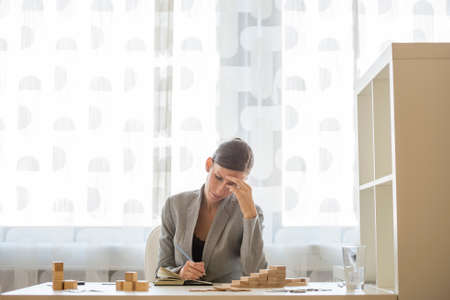 Businesswoman working at office desk writing on notepad beside stacks of wooden blocks on her desk...