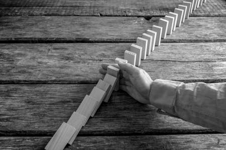 Monochrome image of a businessman preventing dominoes from crumbling with palm on wooden table.