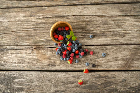 Top view of mixed berry fruits, blueberries, strawberries, blackberries and raspberries,  scattering out of wooden bowl over a textured rustic wooden desk.の写真素材