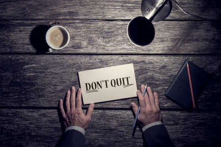 Motivational business concept - Don't Quit with underlined word Do It written on a card lying on a rustic wood desk with a mans hands holding a pen, lamp and mug of coffee viewed from above.の写真素材