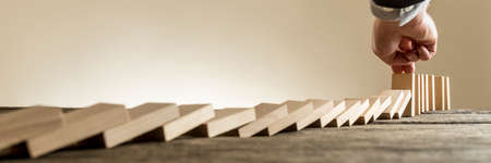 Male hand in business suit stopping domino effect of wooden blocks for concept about strategy and assistance, wide low angle view.