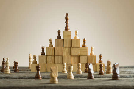 Business hierarchy; ranking and strategy concept with chess pieces standing on a pyramid of wooden building blocks with the king at the top with copy space.