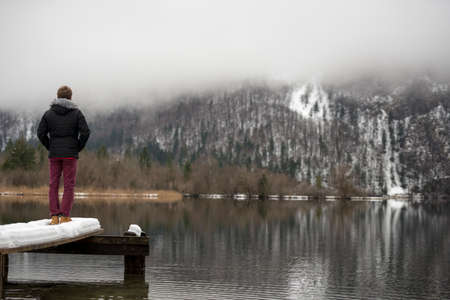 View from behind of a man standing at the end of an old wooden pier covered with snow on a lake Bohinj.の写真素材