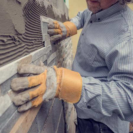 Closeup of manual worker in protection gloves pushing the tile into the cement on the wall while tiling a wall with ornamental tiles, retro effect faded look.の写真素材