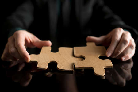 Businessman in black suit holding two matching puzzle pieces, isolated on black background.の写真素材