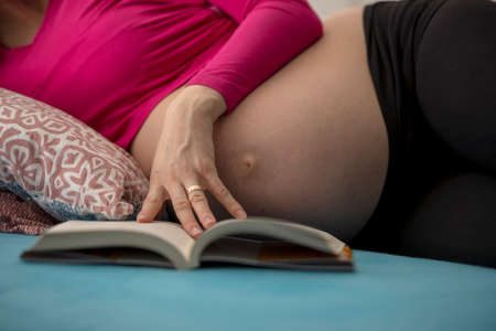 Closeup of a pregnant woman in pink shirt lying on her bed reading a book.の写真素材