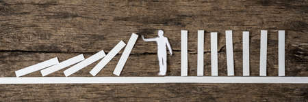 Man stopping the domino effect in a conceptual image of a white paper cutout silhouette on a rustic wood background.の写真素材