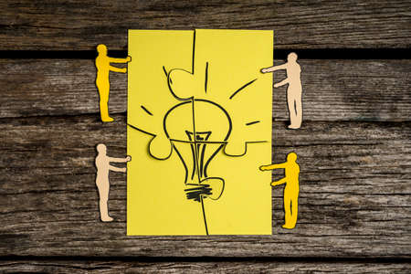 Four paper cutout silhouettes of men holding pieces of a jigsaw puzzle bearing the image of a light bulb to find the perfect idea.の写真素材