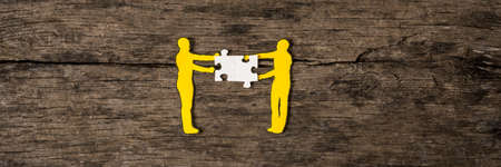 Two yellow paper cutout silhouettes of men holding white puzzle pieces on rustic wood.の写真素材