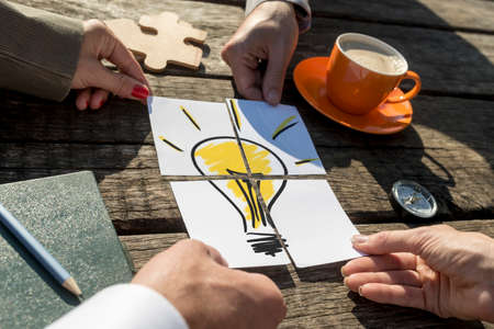 Brainstorming conceptual image of the hands of four business people putting together the pieces of a light bulb on a rustic wooden table outdoors in the sunlight.の写真素材