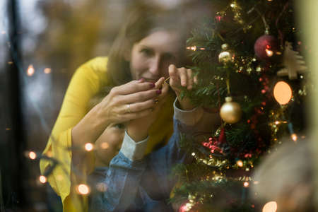 Happy young mother and her toddler son decorating Christmas tree visible behind the window with reflection and copy space.の写真素材