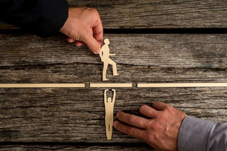 Partnership concept using paper cut outs of men with one holding up a bridge in a gap so that the other can walk across over rustic wooden background.の写真素材