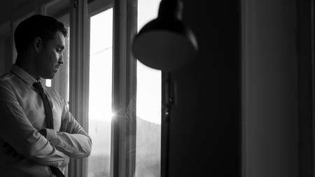 Monochrome image of a young business man wearing white shirt and black tie while thinking and looking through the window at sunset.の写真素材