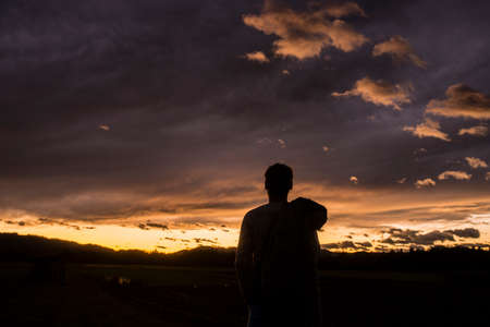 Silhouetted figure of a person against a sunset sky with fiery glow over the horizon and clouds.