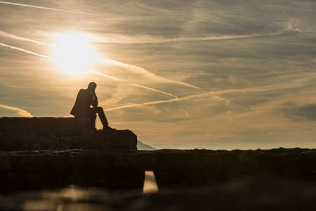 Pensive businessman in elegant suit sitting on a ruins of a castle looking into the distance under a majestic morning sky with a rising sun.の写真素材