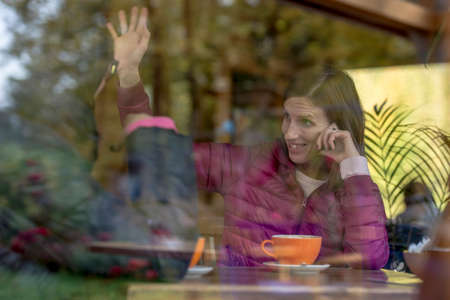View through a glass window with reflections of a young woman sitting indoors at a table smiling and...