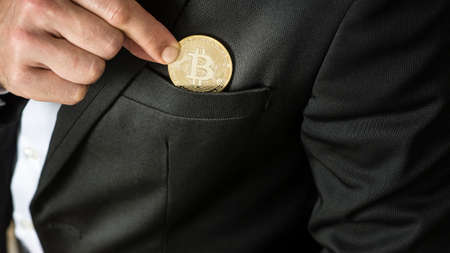 Businessman placing a Bitcoin in his jacket pocket in a close up view on his fingers and the coin.の写真素材