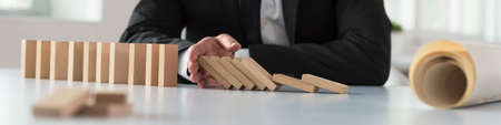 Wide panorama of a businessman stopping the domino effect by inserting his hand into a line of falling wooden blocks with a blueprint rolled alongside.の写真素材