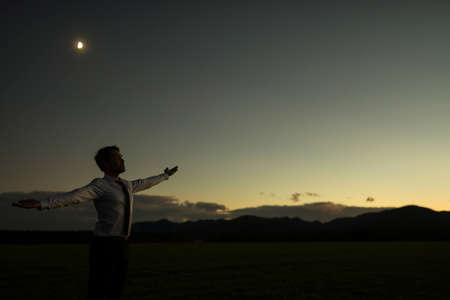 Businessman celebrating a new dawn outdoors in a rural field standing with outspread arms as the glow of the sun appears over the mountains.の写真素材