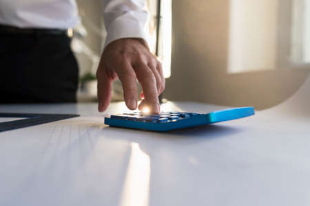 Businessman working on a blue manual calculator in front of a window with right sun flare in a low angle close up view of his hand with copy space.の写真素材