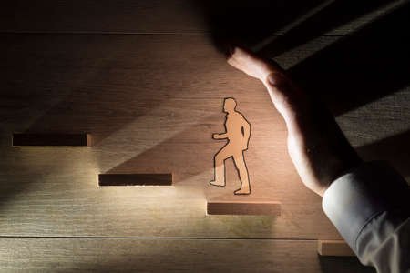 Conceptual close-up of the hand of a man protecting a paper man during progress while climbing stairs against wooden background for copy space.の写真素材