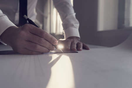 Retro style image of an architect working on blueprint at a table in an office backlit by a bright sunlight behind him in a close up view of his hand and paperwork.の写真素材