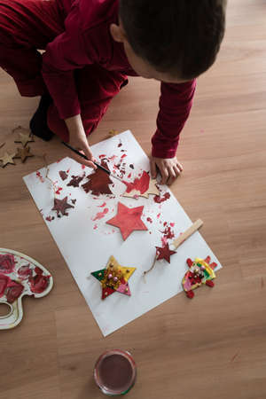Young boy painting rustic homemade Christmas decorations kneeling down on the floor with a box of festive red paint viewed high angle with copy spaceの写真素材