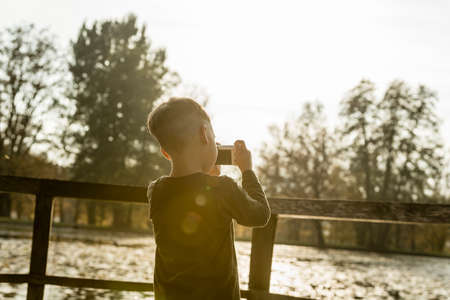 Young boy photographing a lake in a rear view with sun flare and copy space.の写真素材