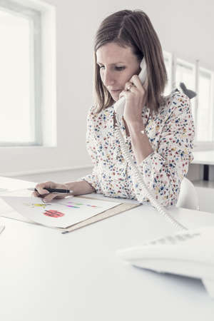 Serious businesswoman making a phone call as she sits at her desk in the office analyzing important data.の写真素材