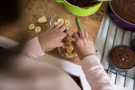 Woman placing sliced banana in a glass bowl while making individual desserts in a high angle over the shoulder view of the hands.の写真素材