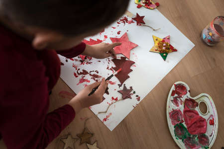 Toddler painting wooden decorations red on a sheet of paper kneeling on the floor.の写真素材