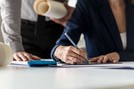 Two architects at work in their office with the man holding a blueprint leaning over a woman drawing using a set square in a close up view of their hands.の写真素材