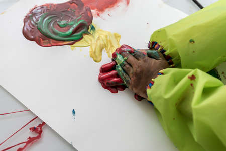 Child wearing protective clothing playing with colorful paints doing hand painting daubing pigment onto paper in an abstract design.の写真素材