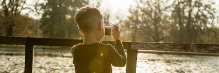Little boy photographing nature standing in front of wooden railings overlooking a lake or river in a panorama rear view with sun flare.の写真素材