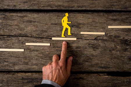 Businessman climbing the steps in a conceptual image with paper silhouette cutouts of a man and a human hand helping him climb over rustic wood.の写真素材
