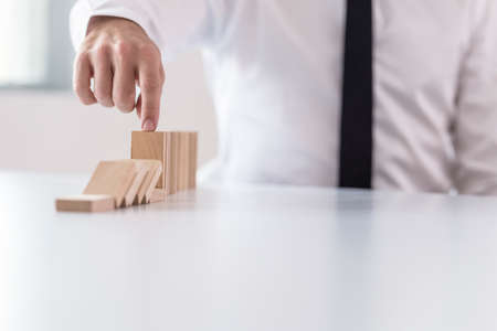 Business man interrupting domino effect by stopping wooden dominoes bricks from crumbling with his finger.の写真素材