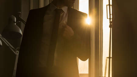 Businessman leaning on a window frame in a shadowy office with a bright sunburst over his shoulder through the window pane.の写真素材