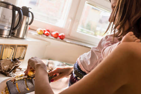 Young mother preparing a meal in the kitchen while her baby clings to her neck in a close up over the shoulder view.の写真素材