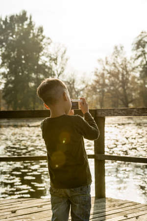 Rear view of a little boy photographing nature with a compact camera outdoors at the lake backlit by sunlight.の写真素材