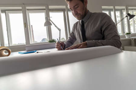 Architect, designer or draughtsman at work in the office on a large blueprint or plan in a cropped low angle view across the table with copy space.の写真素材